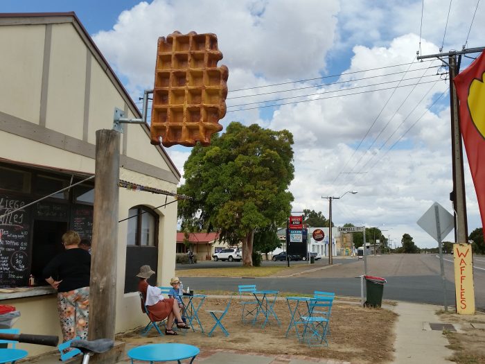 Cafe with blue tables and chairs outside. Woman stands at the window to orer. Woman and child sit at one of the tables. A large waffle is on a pole and another sign that reads 'waffles' is propped against a pole.