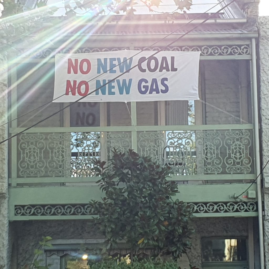 House with sign on balcony that reads "No New Coal. No New Gas". A beam of sunlight crosses the left hand corner.