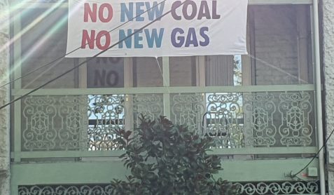 House with sign on balcony that reads "No New Coal. No New Gas". A beam of sunlight crosses the left hand corner.