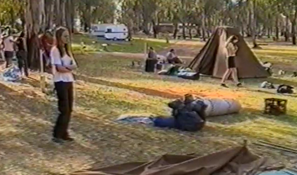 Girl stands alone with arms folded in grassy area with gum trees in the background. There are tents and camping gear scattered around and groups of people in the background.