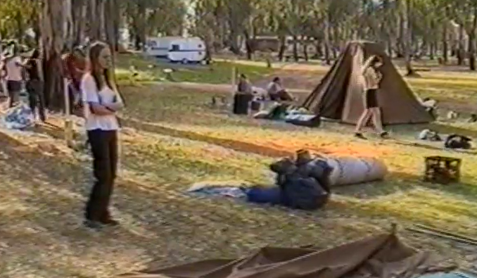 Young girl with long straight hair standing on the grass at a campsite. In the background is a tent and groups of people.